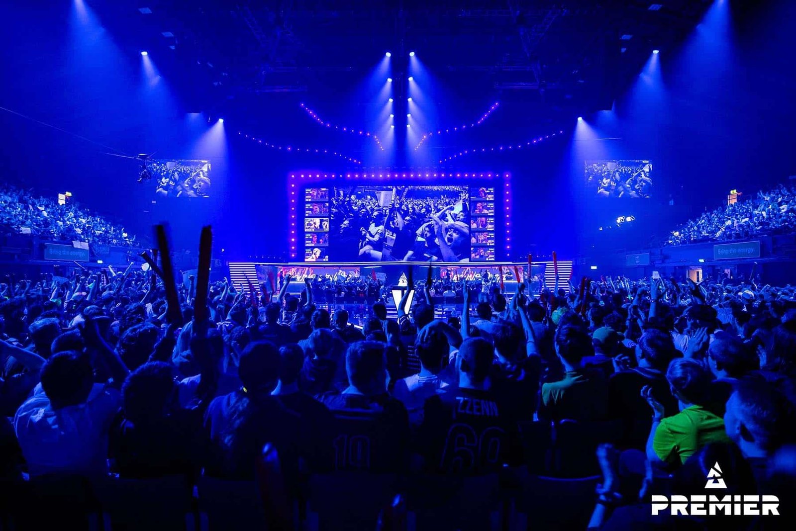 Image of BLAST Premier Spring Final 2024 stage illuminated in blue lights. A crowd is surrounding a podium holding the trophy while Counter-Strike gameplay is displayed on a screen above the stage